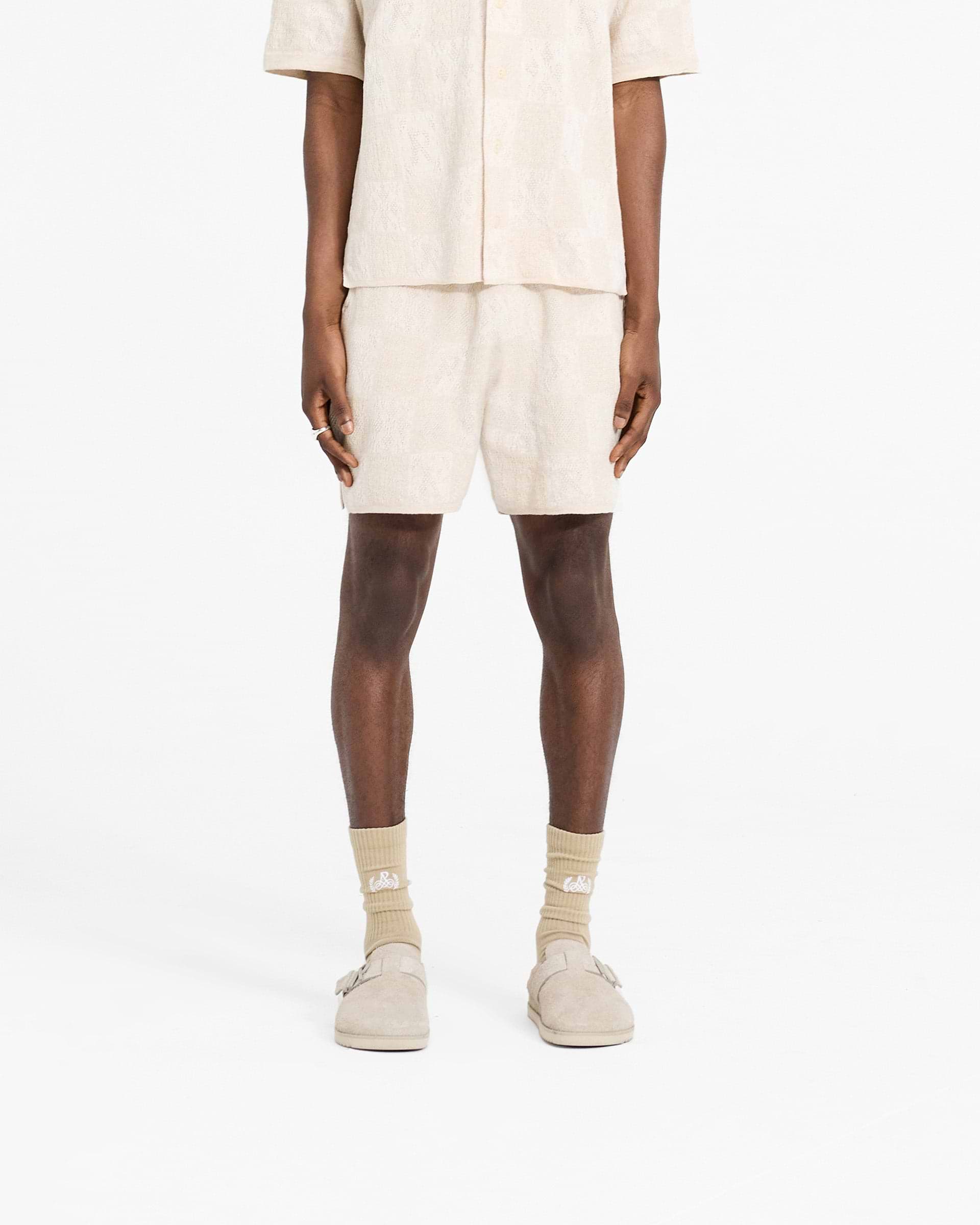 Man wearing cream crochet shorts on a plain white background | Crochet Short in Cream | Drapey crochet knit shorts with intarsia pattern | SC24 Shorts | Represent Clo