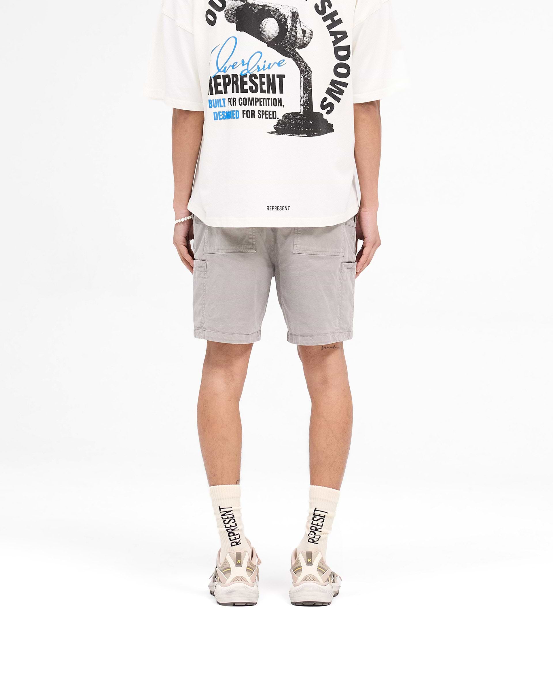 Man wearing mens grey cargo shorts on a plain white background | Season Cargo Short in Washed Taupe | Shorts with welt cargo and diagonal hand pockets | SS25 Shorts | Represent Clo