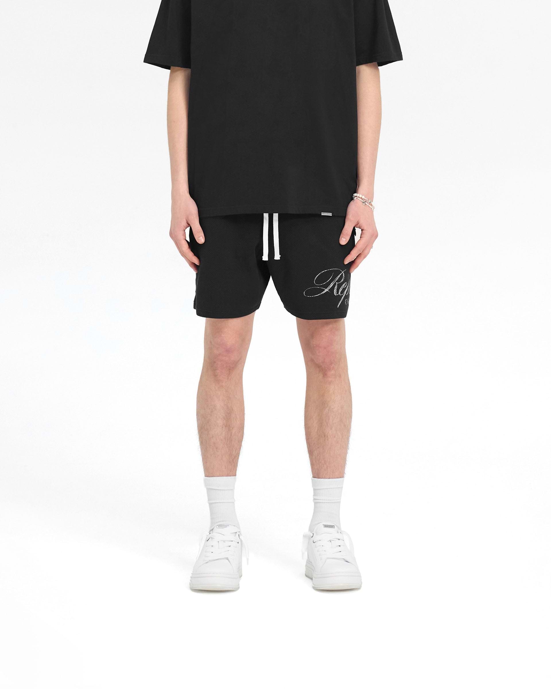 Man wearing black mesh shorts on a plain white background | Represent Owners Club Script Mesh Shorts in Black | Mesh shorts with elasticated waistband and drawcord | Owners Club Shorts | Represent Clo