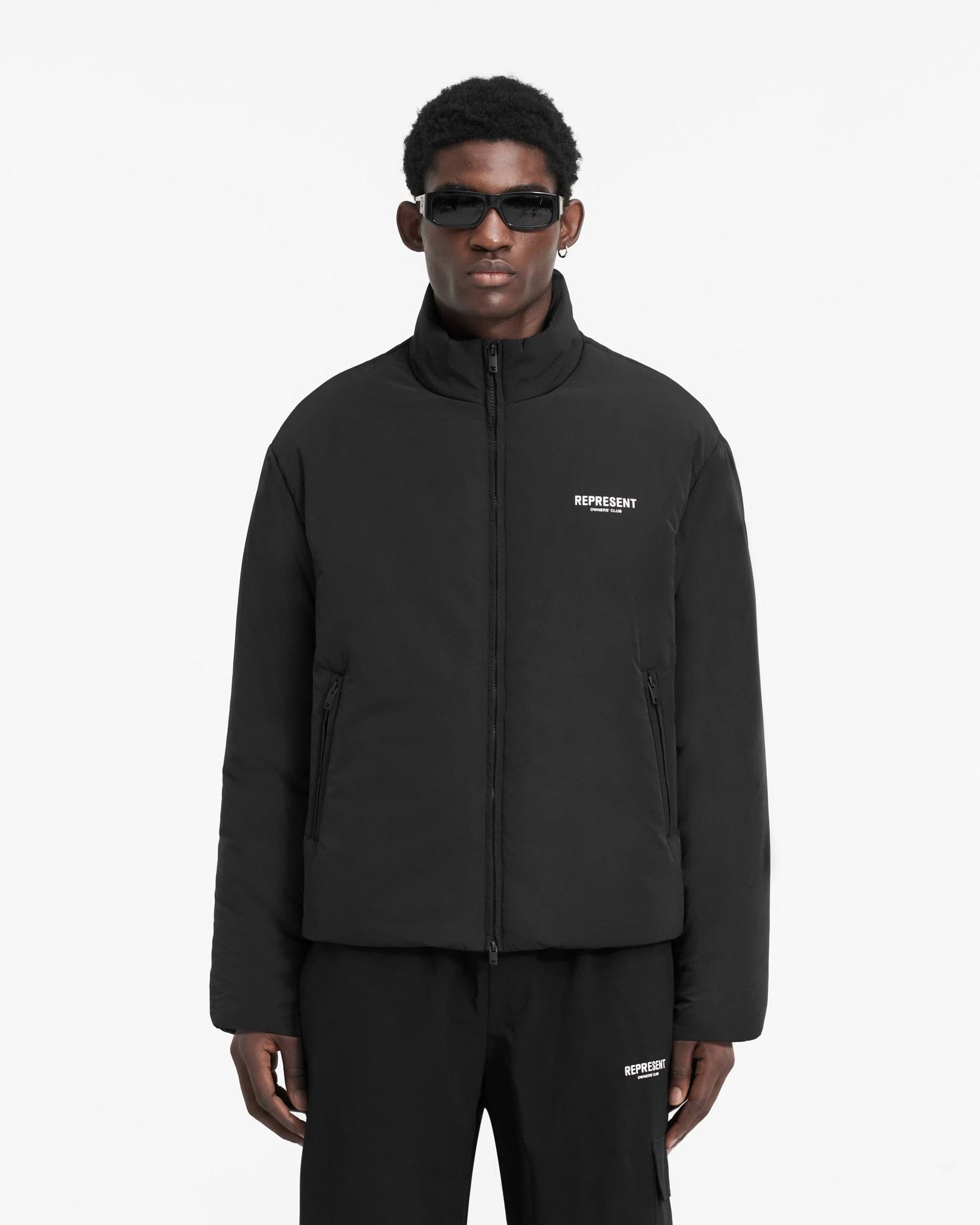 Man wearing a black wadded jacket on a plain white background | Represent Owners Club Wadded Jacket in Black | Down fill jacket with funnel collar and zip pockets | Owners Club Outerwear | Represent Clo
