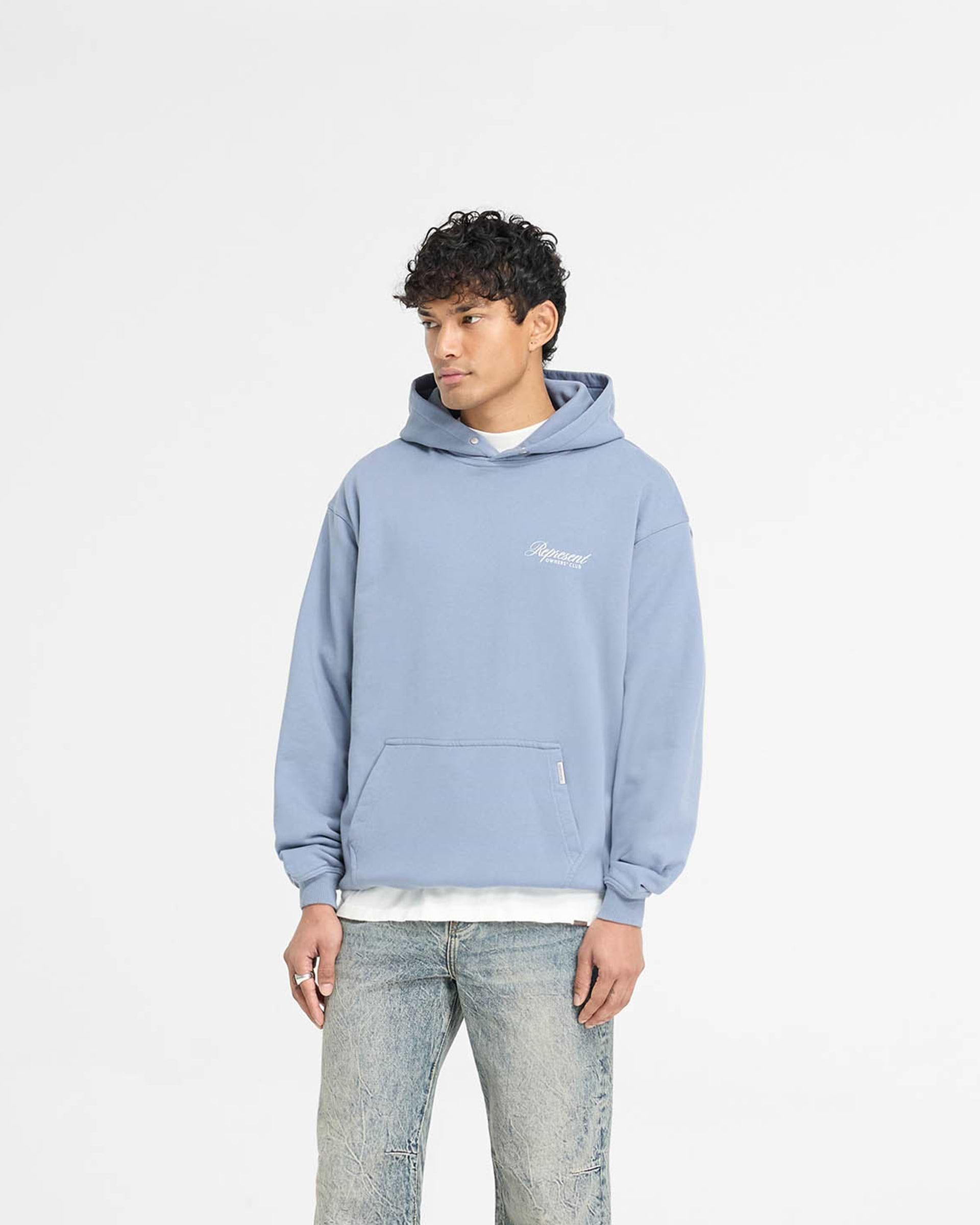 Man wearing a dusty blue hoodie on a plain white background | Represent Owners Club Script Hoodie in Dusty Blue | Heavy weight oversized hoodie with screen print graphic | Owners Club Hoodies | Represent Clo