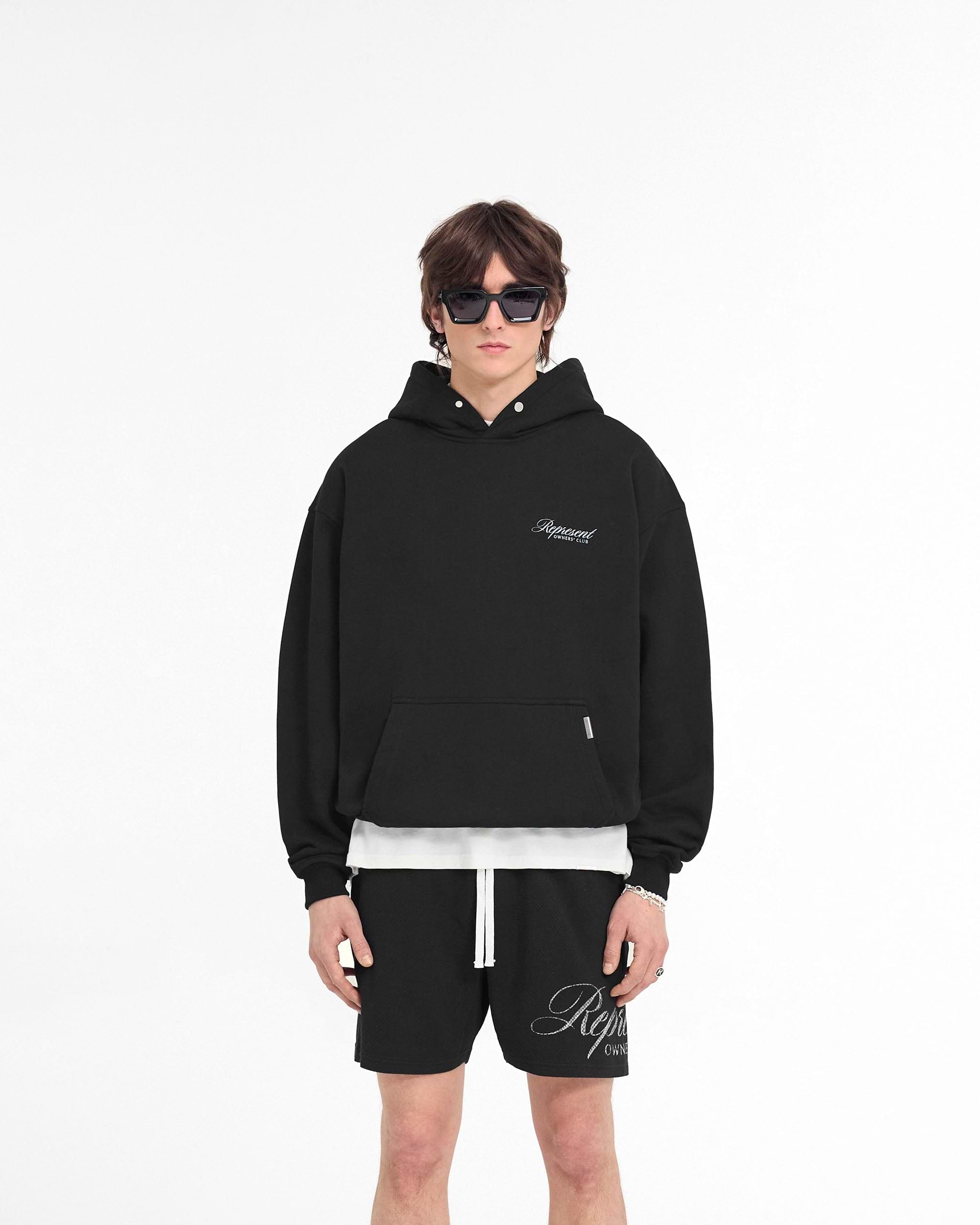 Man wearing a black hoodie on a plain white background | Represent Owners Club Script Hoodie in Black | Oversized fit hoodie with script graphic | Owners Club Hoodies | Represent Clo