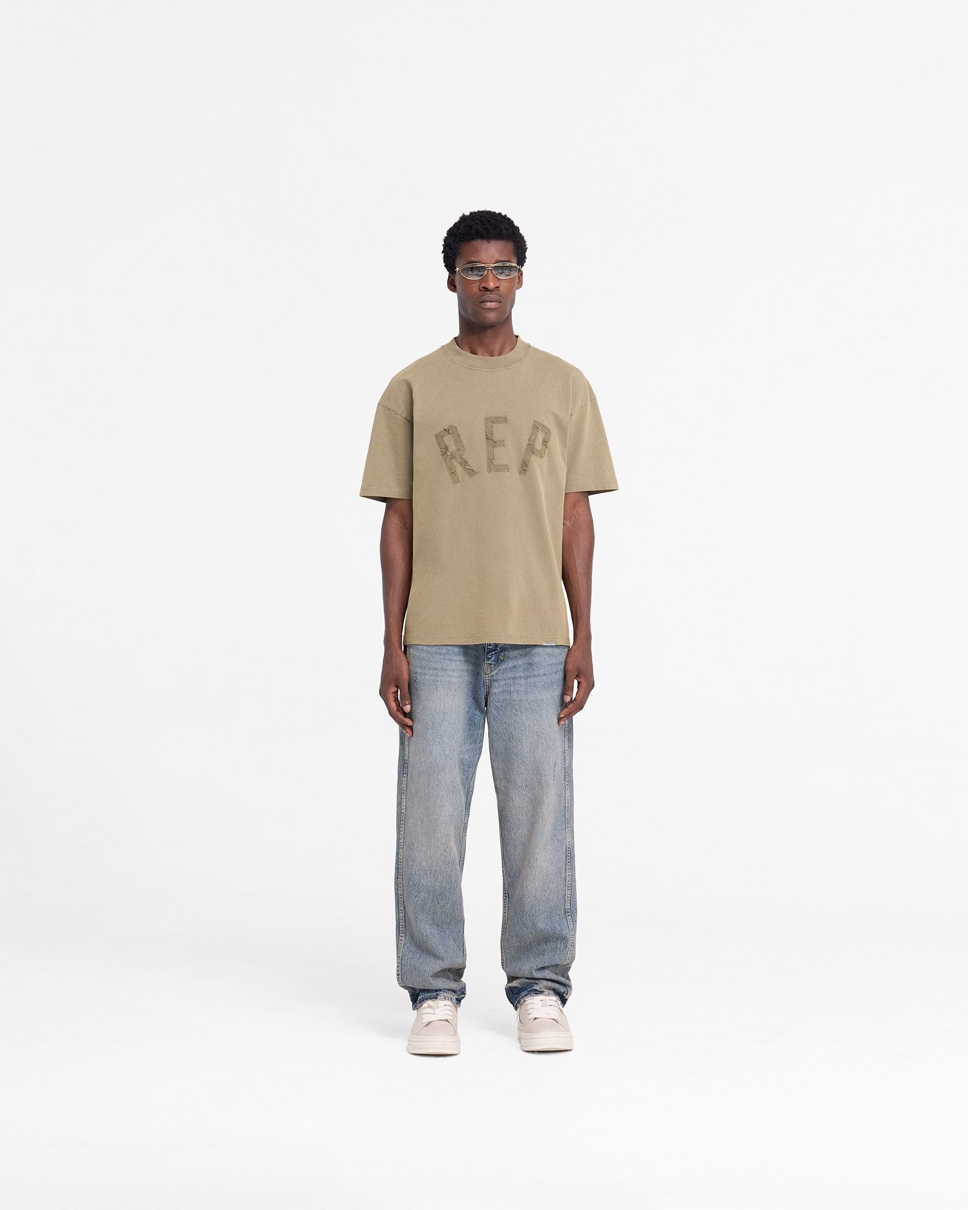 Man wearing a fawn t-shirt on a plain white background | Rep Applique T-Shirt in Fawn | Oversized t-shirt with distressed applique logo | FW24 T-Shirts | Represent Clo