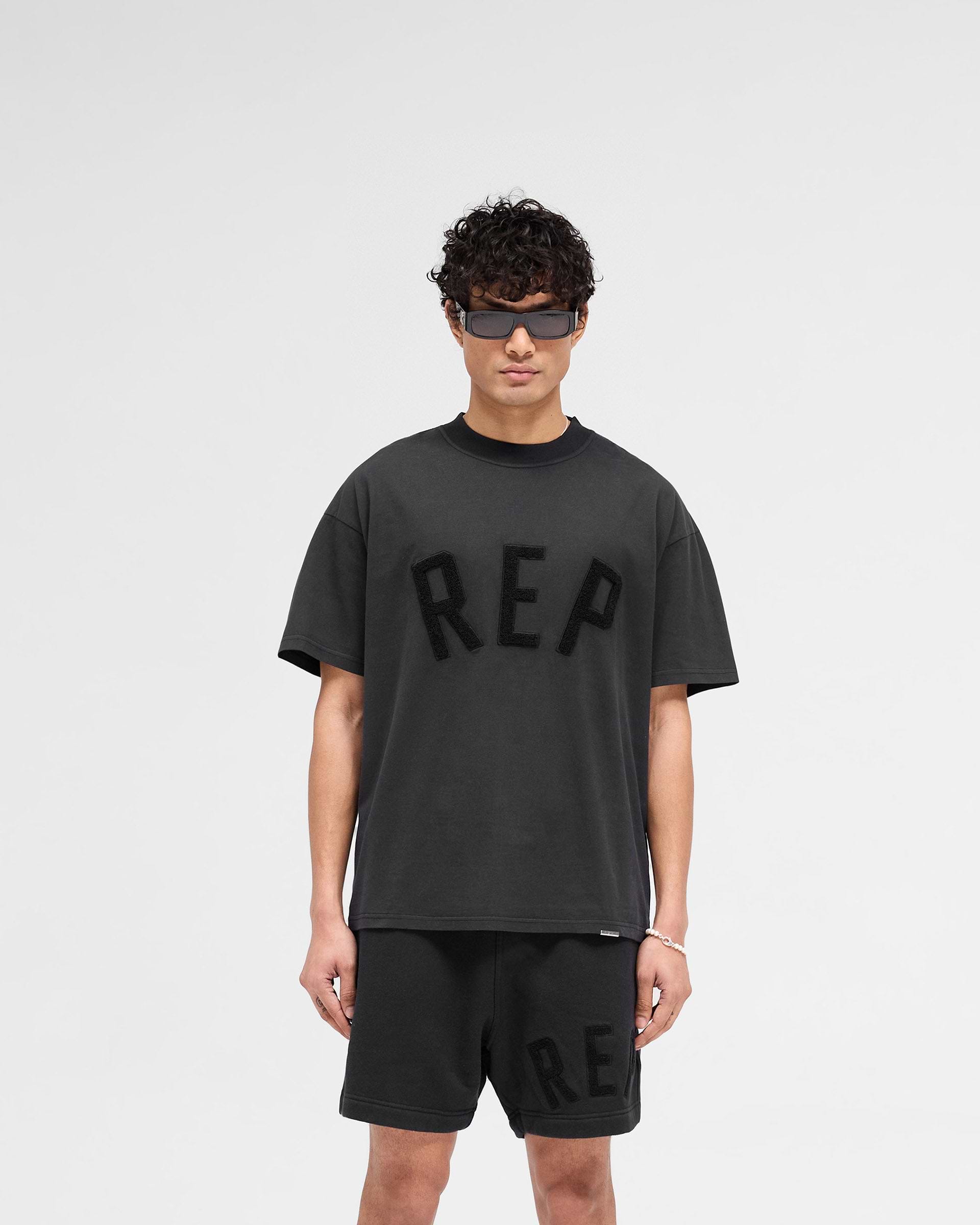Man wearing a slightly oversized black t-shirt on a plain white background | Rep Applique T-Shirt in Off Black | T-shirt with textured applique and overlock seams | SS25 T-Shirts | Represent Clo