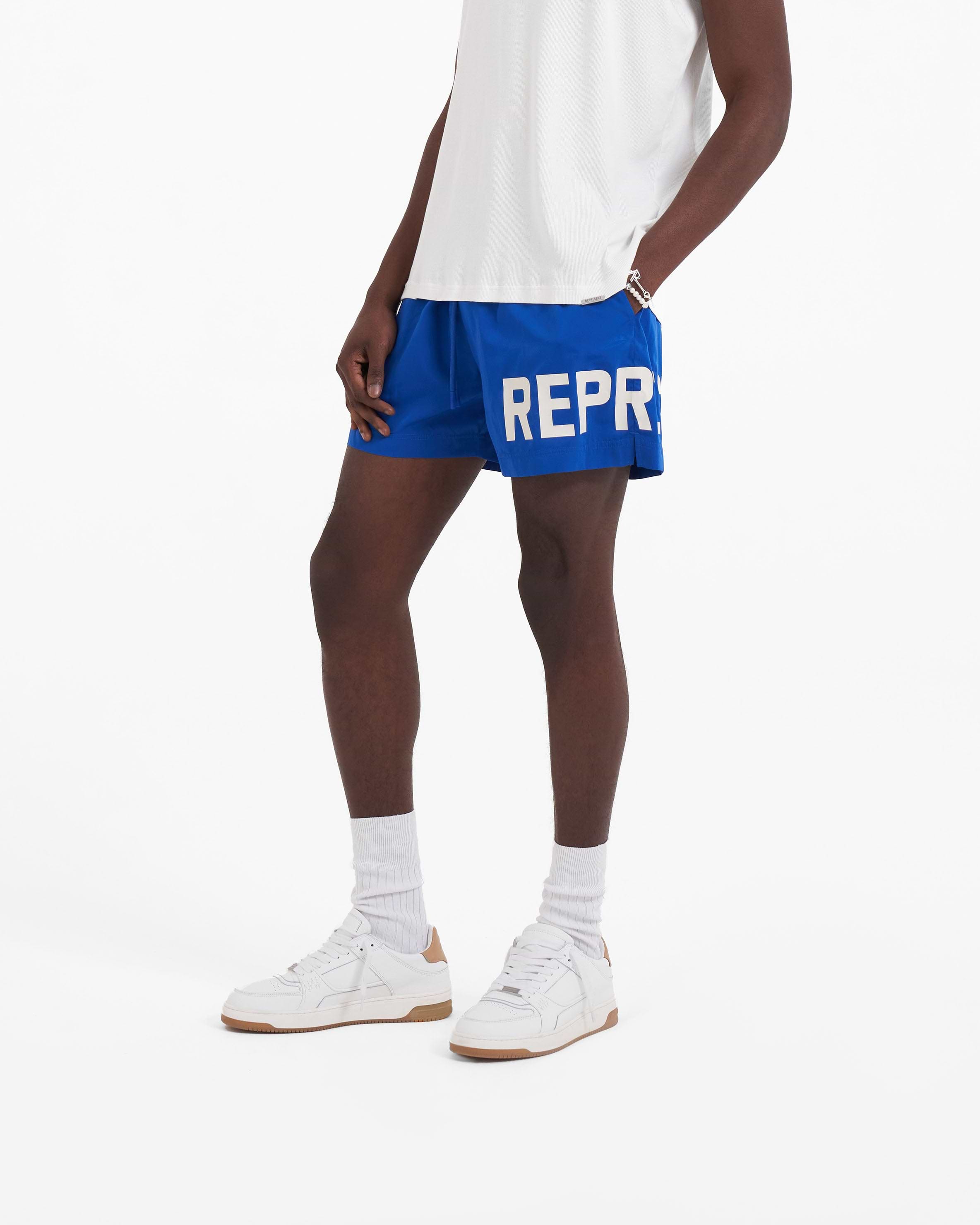 Man wearing cobalt blue swim shorts on a plain white background | Swim Shorts in Cobalt | Shorts with elasticated waistband and logo print | SC23 Shorts | Represent Clo