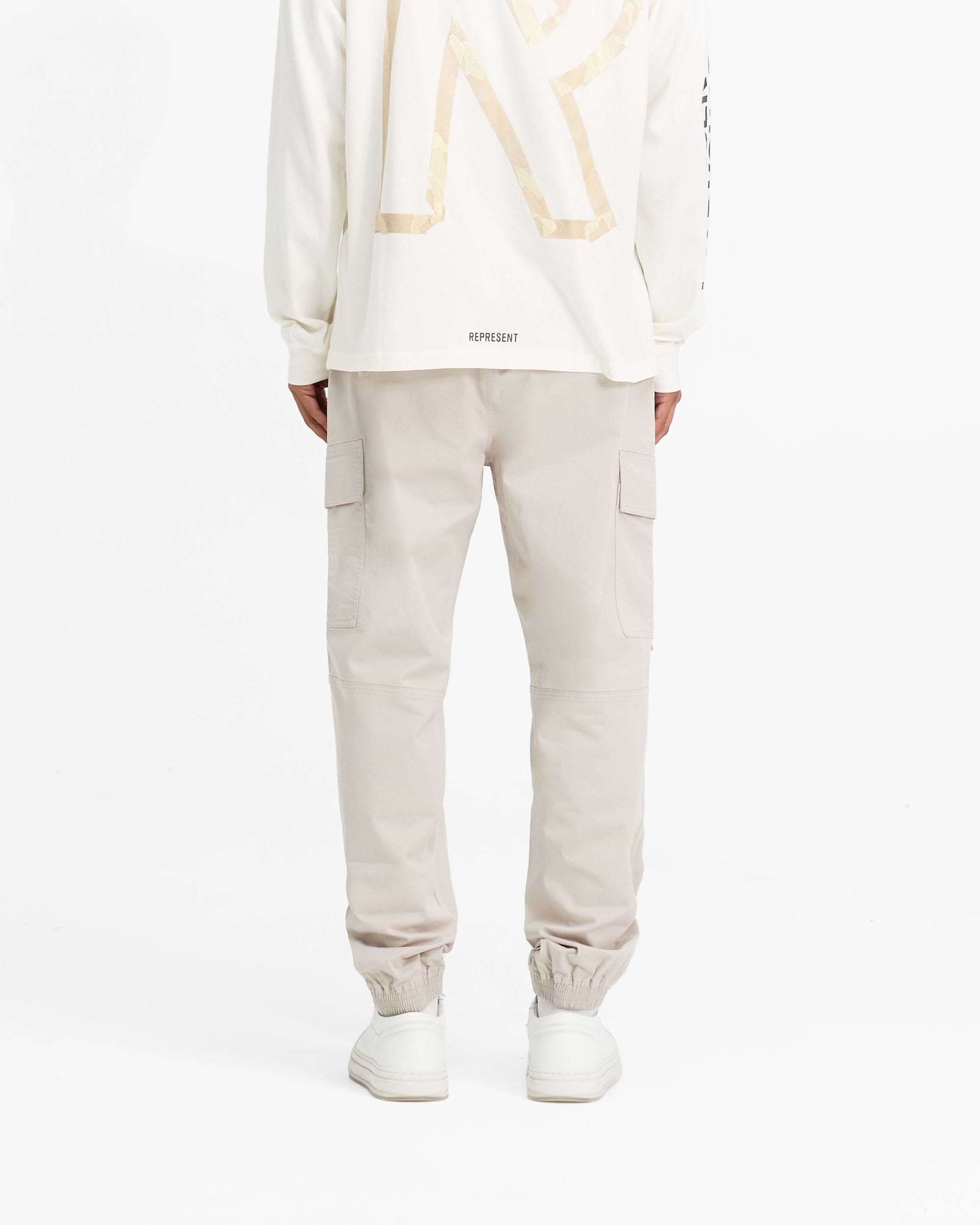 Man wearing stone cargo pants on a plain white background | Cuffed Cargo Pant in Stone | Cargo pants with elasticated cuffs and waistband | FW24 Pants | Represent Clo