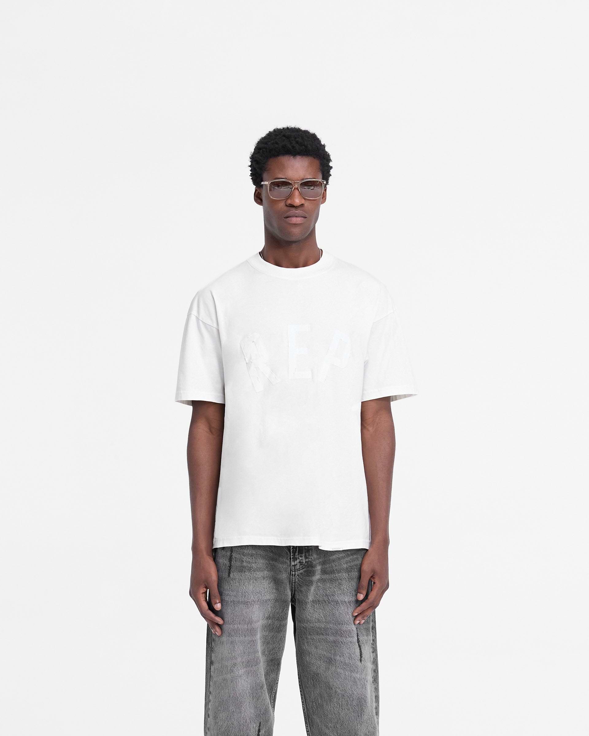 Man wearing a white t-shirt on a plain white background | Rep Applique T-Shirt in Flat White | Oversized t-shirt with distressed applique logo | FW24 T-Shirts | Represent Clo
