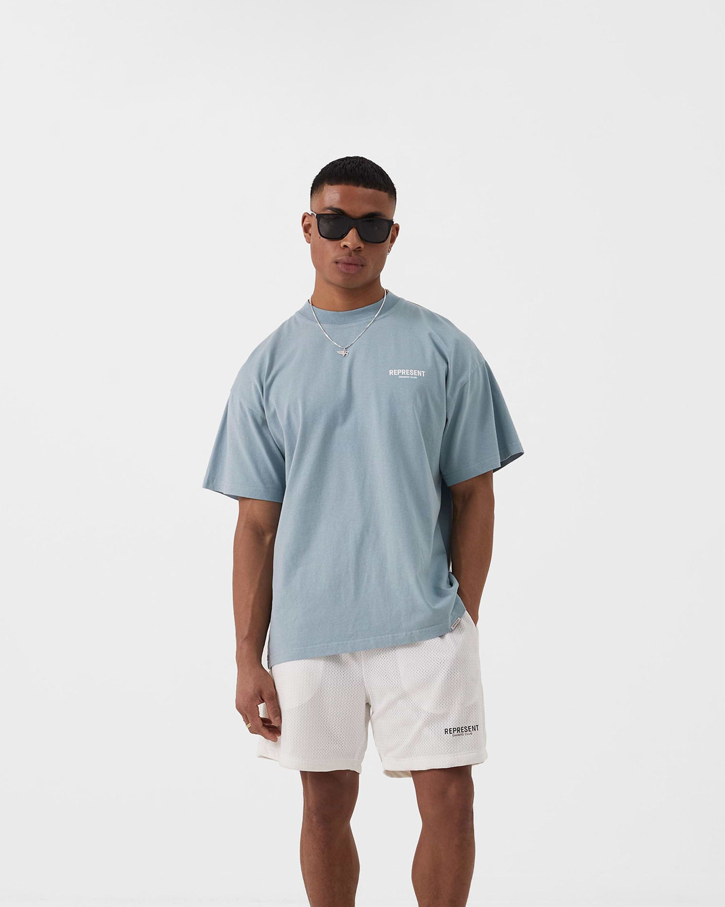 Man wearing a blue t-shirt on a plain white background | Represent Owners Club T-Shirt in Powder Blue | Oversized fit cotton t-shirt with graphic branding | Owners Club T-Shirts | Represent Clo